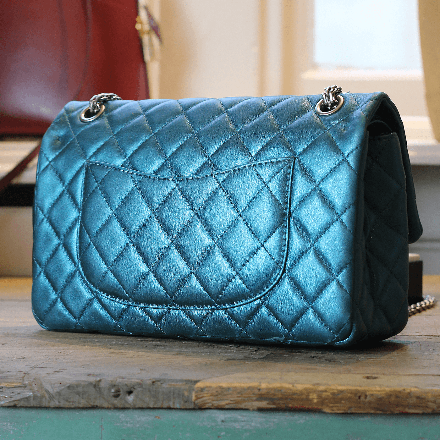The back of a shiny blue quilted Chanel crossbody handbag with a pocket on the outside. The bag is sitting on a wooden antique table inside a vintage shop.