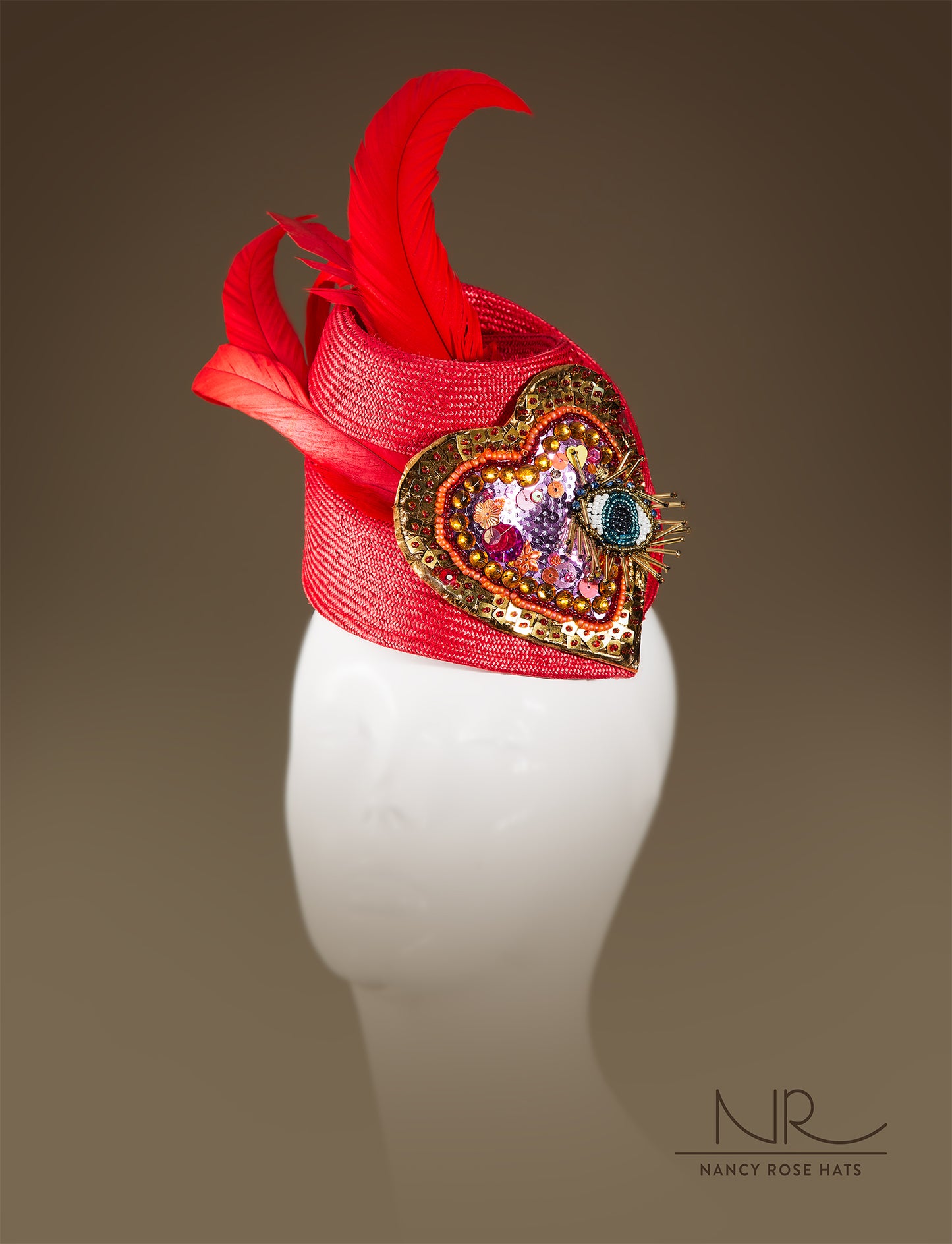 A distinctive red hat made of swirling sinamay with long red feathers and a large heart-shaped embellishment made of sequins and beads with a big eye made of beading in the center, displayed on a mannequin head.